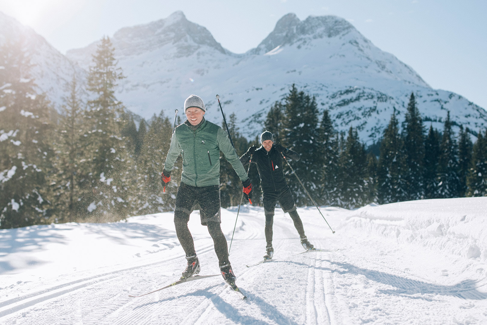 Zwei Langläufer fahren sportlich auf einer sonnigen Loipe durch verschneite Winterlandschaft mit Bergen und Wäldern.