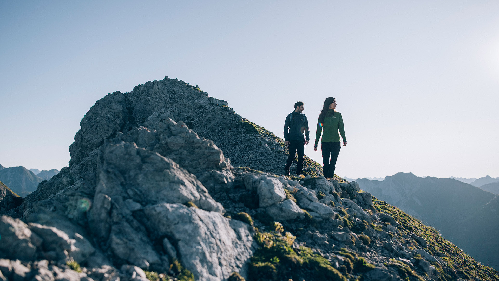 Zwei Wanderer gehen über felsiges Gelände in den Bergen, bei klarer Sicht und tiefstehender Sonne, umgeben von alpinem Panorama.