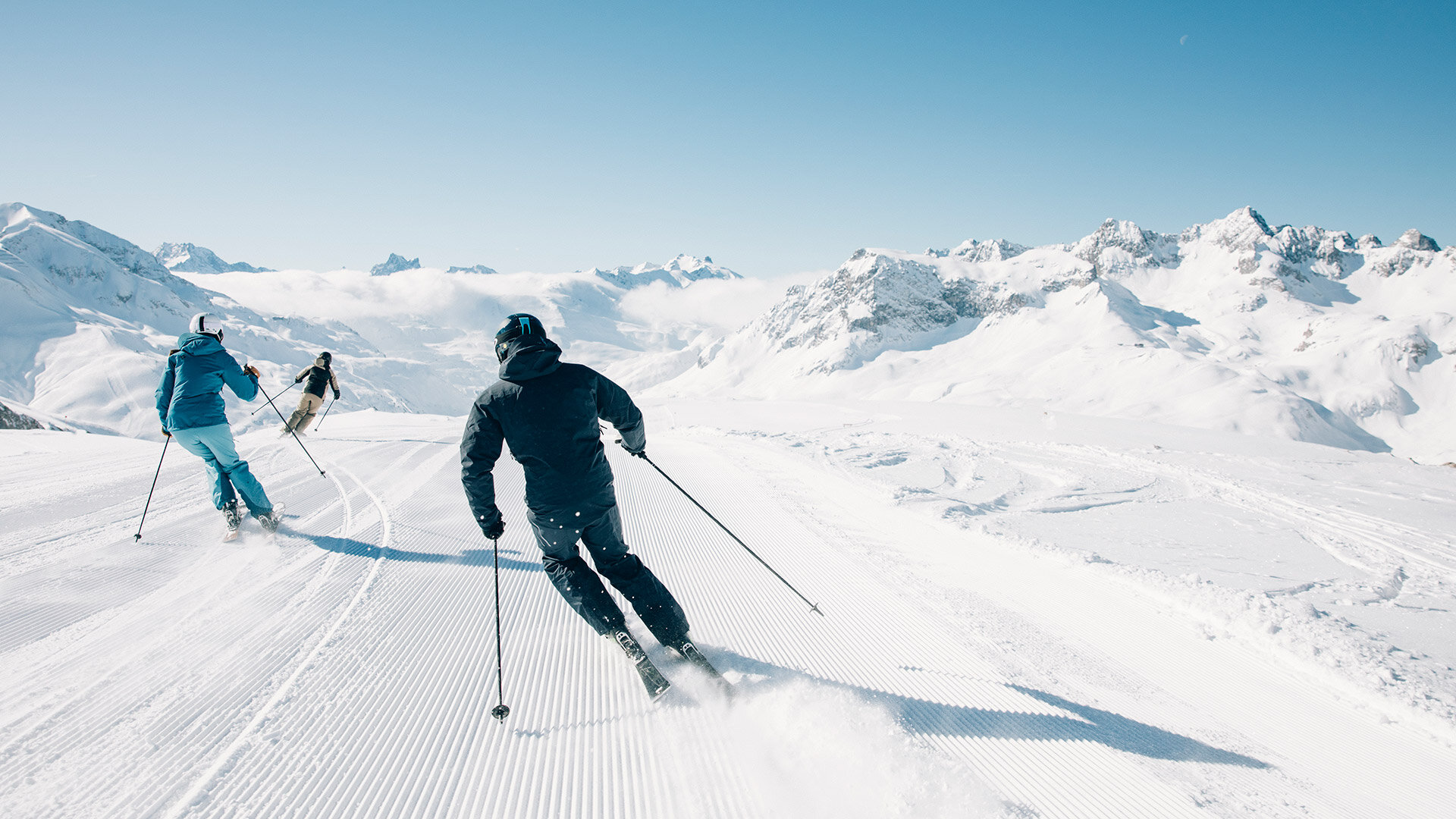 Zwei Skifahrer fahren auf präparierter Piste durch sonnige, verschneite Winterlandschaft mit Bergen und blauem Himmel.