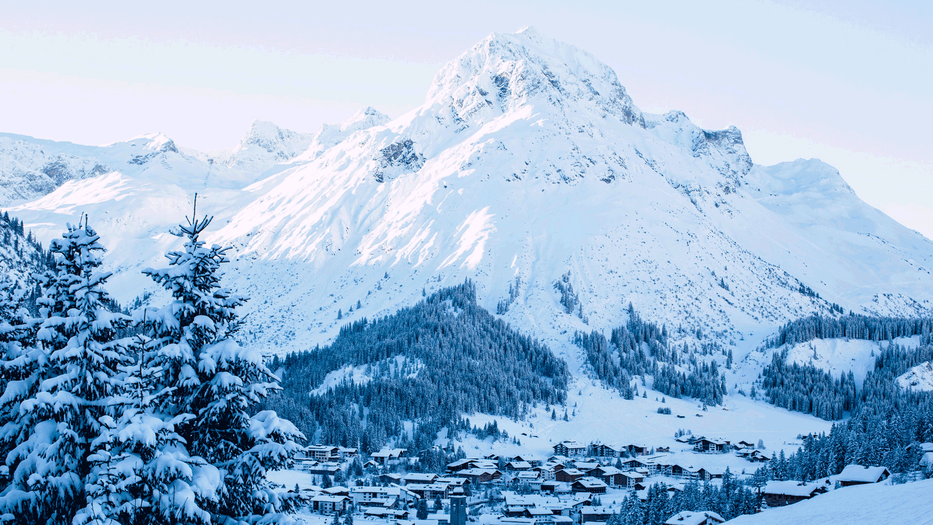 Omeshorn, Lech Verschneite Berglandschaft mit einem Dorf im Tal, schneebedeckte Tannen im Vordergrund und markanter Gipfel im Hintergrund.